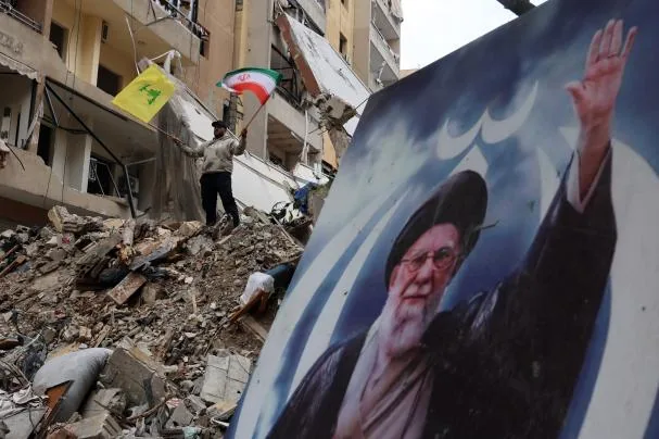 A Hezbollah supporter, on the ruins of a building in Beirut from which a portrait of the late Ali Khamenei protrudes